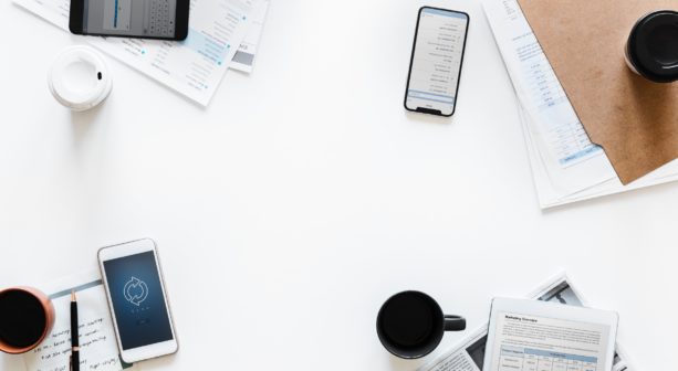 A birds-eye view of a white desk with 3 smartphones, a tablet computer, file folders, and 3 coffee mugs