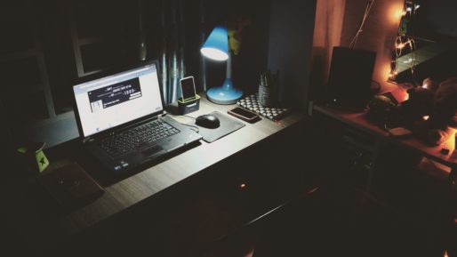 A desk lit by a small, blue desk lamp with a laptop computer and a smartphone