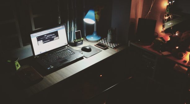A desk lit by a small, blue desk lamp with a laptop computer and a smartphone