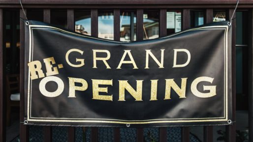 A black banner hanging by grommets with uppercase gold text, reading RE-GRAND OPENING